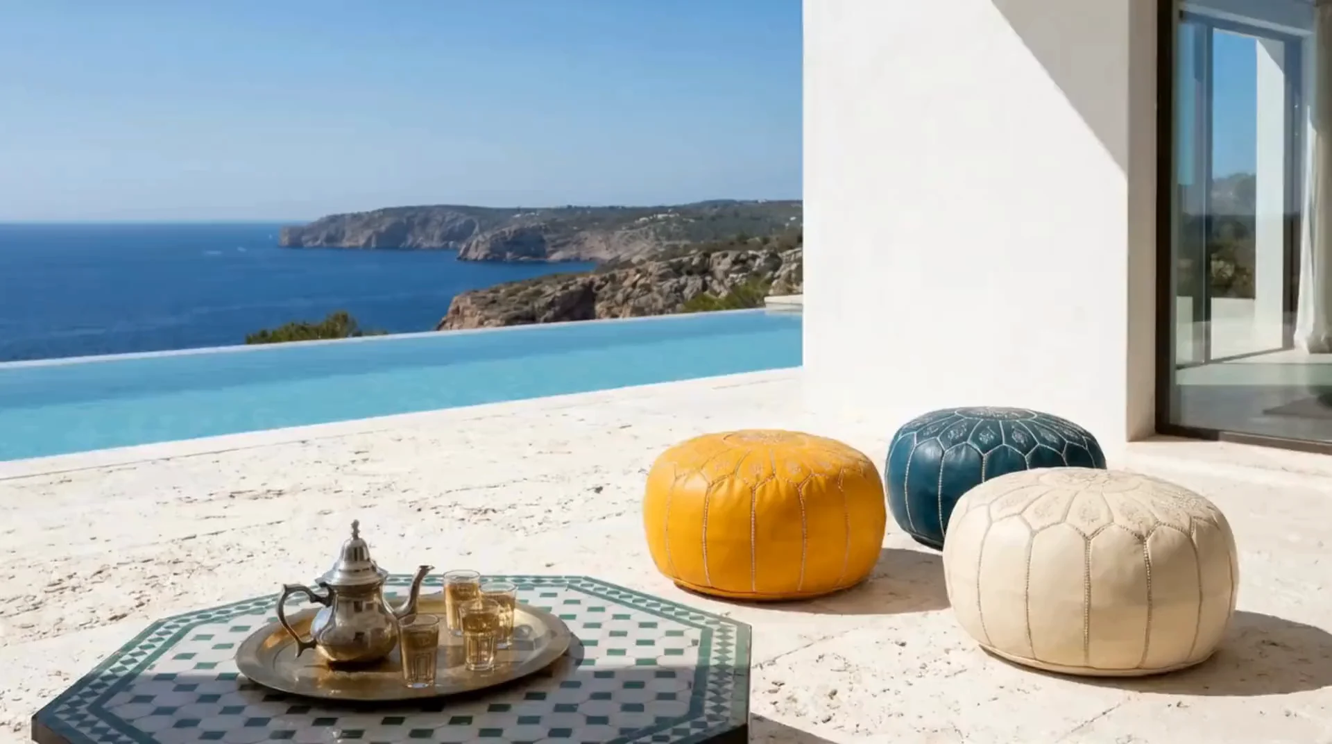 Mustard, teal and ivory Moroccan poufs beside a tea tray on a poolside terrace overlooking the Mediterranean