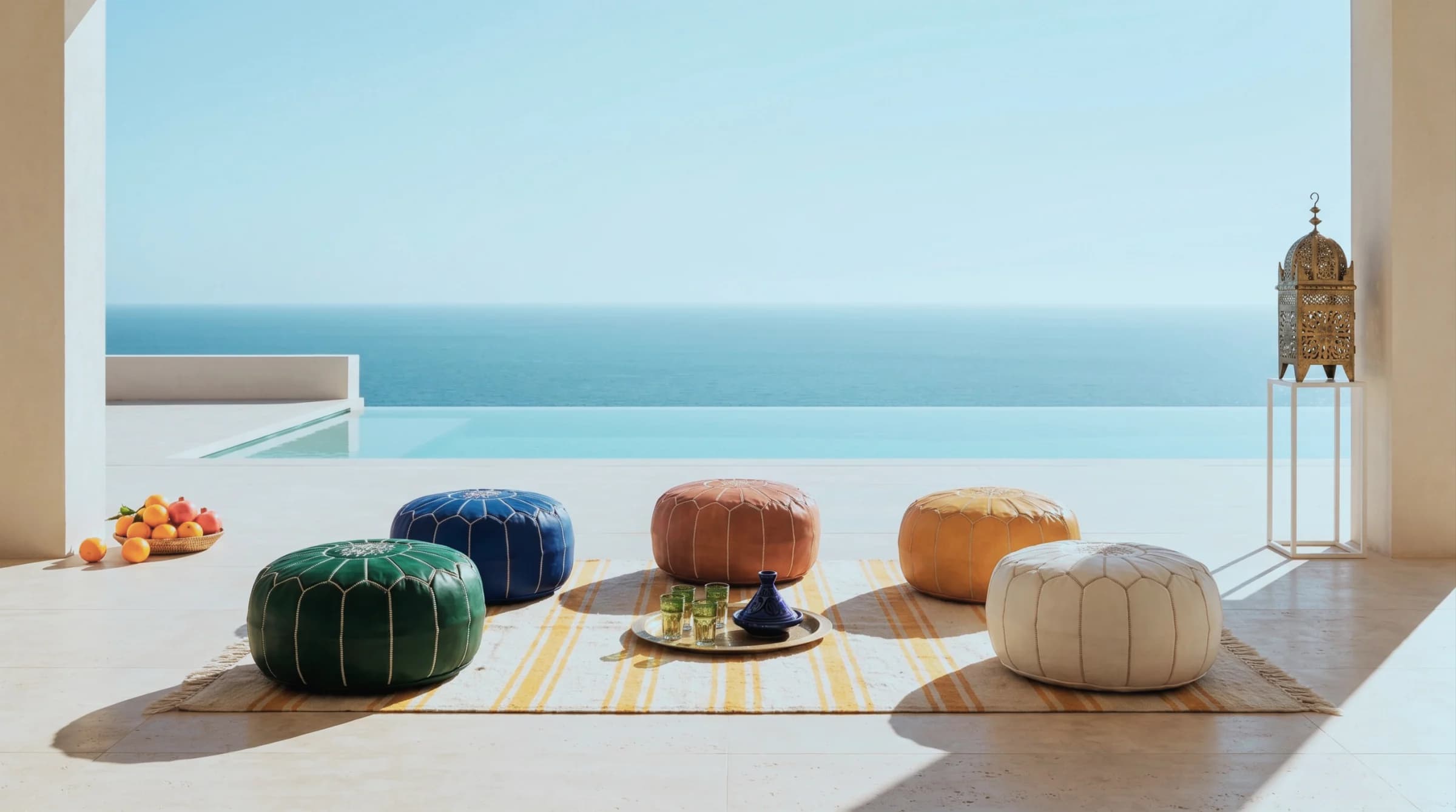 Three Moroccan leather poufs in mustard, teal and ivory grouped around a hand-painted zellige side table on a cliff-top Mediterranean terrace